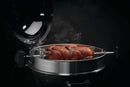 A whole seasoned chicken roasts on a rotisserie inside a Napoleon Grills PRO Charcoal Kettle, using the Napoleon Commercial Grade Rotisserie Kit (69241), with the lid partially open and smoke rising against a dark background.