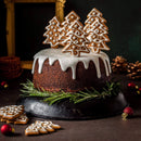 Chocolate cake with white icing, delicately dusted with The Spice Lab Organic Ground Ginger and decorated with gingerbread cookies shaped like Christmas trees, sitting on a black plate and surrounded by rosemary and festive ornaments.