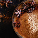 A close-up of The Spice Lab Ground Star Anise pods and powder displayed on a ceramic plate, with some scattered on a dark surface. This aromatic spice from The Spice Lab features a distinctive licorice-like flavor, perfect for adding depth to both sweet and savory dishes.