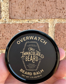 A hand holds a round tin labeled The Immaculate Beard Limited Edition Beard Balm by The Immaculate Beard, featuring nourishing shea butter and the warm scent of sandalwood, against a brick wall background.