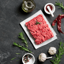 The Dude Food Essentials Meat Box by Dude Food showcased on a dark textured surface, featuring raw ground beef on parchment paper, surrounded by aromatic rosemary sprigs, whole garlic bulbs, peppercorns, vibrant red chilies, a bowl of spice blends, and a bottle of oil.