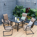 Four brown patio chairs surround a DirectDeals 31 Inch Outdoor Fire Pit Dining Table with Cooking BBQ Grate on a wooden deck, drinks and glasses on top. The space is accented by lush green and colorful plants near a gray slatted fence.