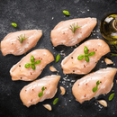 Six boneless, skinless chicken breasts from the Dude Food Chicks Meat Box by Dude Food are hand trimmed for quality and displayed on a dark surface, garnished with herbs, coarse salt, peppercorns, and garlic cloves. A bottle of olive oil appears in the upper right corner.