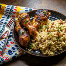 A plate featuring grilled chicken drumsticks garnished with herbs and a dash of The Spice Lab Birdseye Chili Pepper Powder is served alongside rice mixed with black-eyed peas. A colorful patterned napkin and two forks are arranged next to the plate on a wooden table.