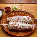 Two elotes (Mexican street corn) on sticks, covered with cheese and a sprinkle of The Spice Lab Organic Chili Powder Blend, served on a decorative brown plate. Lime wedges, extra The Spice Lab Organic Chili Powder Blend, and shredded cheese are in small bowls nearby on a wooden table.
