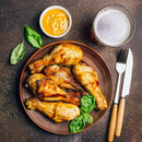 A plate of roasted chicken drumsticks seasoned with The Spice Lab Ground Yellow Mustard 1 lb and foodservice spices, garnished with fresh basil, served with a small bowl of mustard sauce, a glass of beer, and a fork and knife on a dark textured surface.