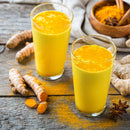Two glasses of vibrant yellow turmeric tea made with The Spice Lab Golden Milk Turmeric Tea Superfood Premium Powder (4 oz) sit on a wooden table, surrounded by fresh turmeric roots, superfood powder from The Spice Lab, star anise, and ginger.