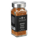 A clear glass jar of The Spice Lab Smoked Ghost Pepper Sea Salt, topped with a silver lid and adorned with a black label, contains coarse reddish-brown salt made with bhut jolokia for those seeking bold flavor and intense heat.