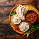 An Indian meal on a woven tray featuring naan bread, bowls of white rice, a bowl of curry seasoned with The Spice Lab Ground Cumin 1 lb Bulk Foodservice Spice, red chili peppers, and fresh cilantro, all presented on a rustic wooden table.