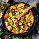 A casserole dish filled with cheesy bread pudding, spinach, and herbs is subtly enhanced with The Spice Lab Ground Yellow Mustard 1 lb by The Spice Lab, and a wooden spatula rests inside. Fresh parsley, a bowl of peppercorns, and a gray napkin are arranged nearby on a dark wooden table.