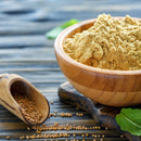 A wooden bowl filled with mustard powder sits beside a small scoop containing The Spice Lab Organic Whole Mustard Seeds by The Spice Lab, arranged on a rustic wooden table—perfect for pickling or brining—with fresh green leaves in the background.