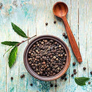 A wooden bowl brimming with The Spice Lab Organic Black Tellicherry Peppercorns Whole is set on a rustic blue wooden surface, accompanied by a wooden spoon and a sprinkling of green leaves.