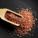 A wooden scoop filled with The Spice Lab Hawaiian Red Alaea Sea Salt Medium Grain spills coarse, reddish-brown crystals onto a dark surface, suggesting their distinctive earthy flavor.