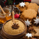 A wooden bowl of brown sugar topped with The Spice Lab Ground Star Anise, surrounded by honey, decorated cookies, star-shaped cookies with white icing, and holiday ornaments on a festive table. This aromatic spice from The Spice Lab infuses the scene with a warm, licorice-like flavor—perfect for celebrating the season.