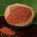 A coconut shell bowl holds The Spice Lab Hawaiian Red Alaea Sea Salt Medium Grain, an earthy-flavored salt from The Spice Lab, with some of the salt spilled beside it. The bowl rests on a dark surface with a green leaf in the background.