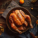 A whole roasted chicken sits in a rustic bowl on a wooden table, garnished with herbs and a sprinkle of The Spice Lab Organic Ground Black Pepper. Nearby are oranges, dried leaves, and a cloth napkin, creating a warm, autumnal setting.