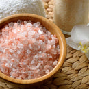A glass grinder from The Spice Lab Himalayan Pink Salt & Premium Black Pepper Grinder Set stands on a woven mat, accompanied by a white towel, a jar, and a white orchid flower.