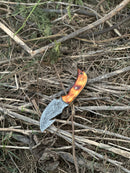 A Texan Knives Custom Handmade Damascus Steel Guthook Skinning Knife with a 7.5” blade and camel bone handle rests outdoors on dry twigs and grass, shown alongside its leather sheath.