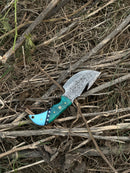 The Texan Knives Elite Custom Handmade Damascus Steel Guthook Skinning Knife features a 7.5” premium blade and a resin handle, displayed on a bed of dry twigs and grass, and comes with a leather sheath.