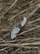 The Texan Knives Custom Handmade Damascus Steel Guthook Skinning Knife with a 7.5” rosewood & resin handle and leather sheath rests on a pile of dry, tangled branches and twigs outdoors.