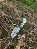 The Texan Knives Custom Handmade Damascus Steel Guthook Skinning Knife, 7.5” with a patterned resin handle and included leather sheath, rests on a bed of dry branches and leaves outdoors.