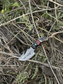 The Texan Knives Premium Custom Handmade Damascus Steel Guthook Skinning Knife features a 7.5" sharp, patterned blade and a vibrant resin handle, shown outdoors among dry twigs and green plants with its leather sheath.