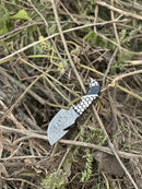 The Texan Knives Exquisite Custom Handmade Damascus Steel Guthook Skinning Knife with a unique resin handle and 7.5” blade rests on dry twigs and green foliage outdoors.