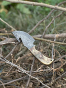 A Texan Knives Custom Handmade Damascus Steel Guthook Skinning Knife with a sheep horn handle and 7.5" blade rests on dry branches outdoors, with a leather sheath included and blurred greenery in the background.