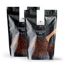 Two black bags of The Spice Lab Ancho Chili + Coffee Rub are displayed, each labeled with the brand name. Perfect for adding rich, smoky flavor to your dishes, this bold BBQ seasoning also works well as a bulk foodservice rub. Beside the bags on a white background lies a wooden spoon filled with the spice blend.