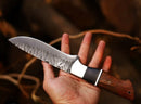 A person holds the Texan Knives 10" J2 hand-forged steel hunting knife with a rosewood handle and leather sheath—a top pick among the best knives for hunting—set against a blurred outdoor background.