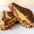 A Reuben sandwich on marbled rye with corned beef, melted cheese, sauerkraut, and Terrapin Ridge Farms Garlic Kraut Mustard, cut in half and stacked on a white plate.