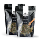 Two black resealable bags of The Spice Lab Chimichurri Seasoning are displayed, one positioned slightly in front, accompanied by a wooden spoon filled with the vibrant Argentine herb blend featured on the packaging.