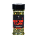 A clear plastic jar of The Spice Lab Chimichurri Seasoning features a black lid and a black label with red and white text. Inside, the jar holds vibrant green and yellow herb seasoning inspired by traditional Argentine chimichurri.