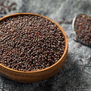 A wooden bowl filled with The Spice Lab Brown Mustard Seeds Whole rests on a gray textured cloth, accompanied by a metal spoon containing more seeds from The Spice Lab in the background—ideal for making homemade mustard or adding bold flavor to your pickling recipes.