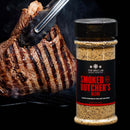 A pair of tongs holds a grilled steak with char marks beside a jar of The Spice Lab Barbecue Seasoning Collection blend—a must-have from The Spice Lab for any serious BBQ enthusiast.