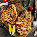 Grilled pork chops on a wooden cutting board, one sliced and garnished with rosemary. Enhanced with The Spice Lab Cooking Essentials Seasoning Set from The Spice Lab, these chops are paired with lemon wedges, cherry tomatoes, garlic, and surrounded by utensils.