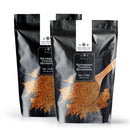 Two black pouches of The Spice Lab Rotisserie Chicken Seasoning stand upright on a white background, each featuring an image of a wooden spoon with seasoning spilling out. The packaging emphasizes the seasoning’s rich golden color and enticing roasted chicken flavor that The Spice Lab Rotisserie Chicken Seasoning brings to your dishes.