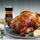 A roasted chicken with a golden color sits on a plate, garnished with lemon slices and rosemary, while a bottle of The Spice Lab Rotisserie Chicken Seasoning in the background hints at its rich, savory flavor.