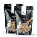 Two black pouches of The Spice Lab Sweet Basil & Garlic Seasoning, each featuring a clear window revealing the flavorful basil and dried spice blend inside, are pictured next to a wooden spoon filled with the savory garlic seasoning mix.