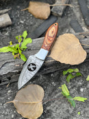The Texan Knives 8" Forged Damascus Skinner Knife with a wire-cut blade, pakkawood & steel bolster handle, and leather sheath rests on a log among dry leaves and green sprouts.