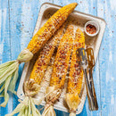 Four grilled corn cobs with husks, sprinkled with grated cheese and a hint of The Spice Lab Organic Chili Powder Blend, on a white tray with metal tongs and a small dish of chili flakes, set on a bright blue wooden surface.