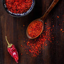 A dried red chili pepper, a spoon, and a bowl filled with crushed red chili flakes are arranged on a dark wooden surface, with some chili flakes scattered around. The vibrant hues reflect the smoky-sweet flavor of The Spice Lab Organic Smoked Paprika—an essential ingredient from The Spice Lab for elevating BBQ rubs or bringing rich depth to your favorite recipes.