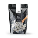 A black resealable pouch labeled “The Spice Lab Salt, Pepper + Garlic Seasoning” from The Spice Lab stands behind a wooden spoon scooping the coarse everyday cooking blend, with some of the seasoning spilling in front of the bag.