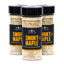 Three jars of The Spice Lab Smoky Maple Seasoning, produced by The Spice Lab, are arranged side by side. Each jar features a black label with yellow text and is topped with a black cap. Inside the jars, the sweet and savory seasoning has a coarse texture and displays a variety of colors that suggest its rich flavor blend.