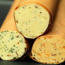 Three logs of compound butter, each wrapped in brown parchment paper and blended with a unique mix of herbs and spices—such as The Spice Lab Lemon Pepper + Thyme Seasoning—are displayed on a dark surface.