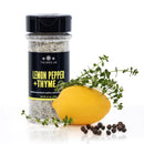 A bottle of The Spice Lab Lemon Pepper + Thyme Seasoning stands beside a fresh lemon, thyme sprigs, and black peppercorns on a white background, offering the perfect citrus herb blend from The Spice Lab to elevate chicken dishes and more.