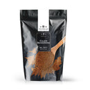A black resealable bag labeled The Spice Lab Cajun Seasoning sits beside a wooden spoon brimming with the vibrant blend, some of the seasoning spilled nearby. The bag displays The Spice Lab’s branding and Net Wt. 2 Lb., offering an authentic Louisiana-style flavor with a mild kick in every sprinkle.