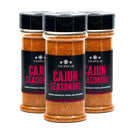 Three bottles of The Spice Lab Cajun Seasoning are grouped together. The transparent bottles reveal vibrant reddish-orange spices inside, each topped with a black lid and featuring a black and red label displaying The Spice Lab brand and product information, promising authentic Louisiana-style flavor.