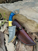 The Texan Knives Skinning Knife features an 8.5" Damascus steel blade, a blue and yellow pakkawood handle with brass bolster, and comes with a button-flap brown leather sheath, shown resting on dry leaves near a rock.