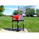 A WPPO LLC Direct Traditional 25" Dual Fueled Pizza Oven With Gas Attachment—featuring both wood and gas power—stands on a black metal wheeled base with a silver chimney, situated on a concrete patio in the middle of a large green grassy field beneath a sunny blue sky.
